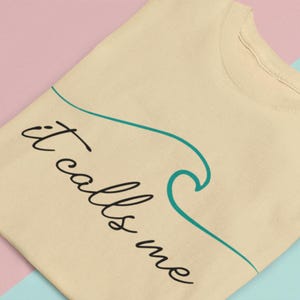 May include: A beige t-shirt with the text "it calls me" in black cursive writing. A teal blue wave graphic is drawn next to the text.