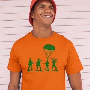 Peut inclure: Un homme portant un t-shirt orange avec une silhouette verte de quatre soldats jouets et un parachute. Les soldats tiennent des fusils jouets et le parachute est au-dessus du soldat &agrave; l'arri&egrave;re.