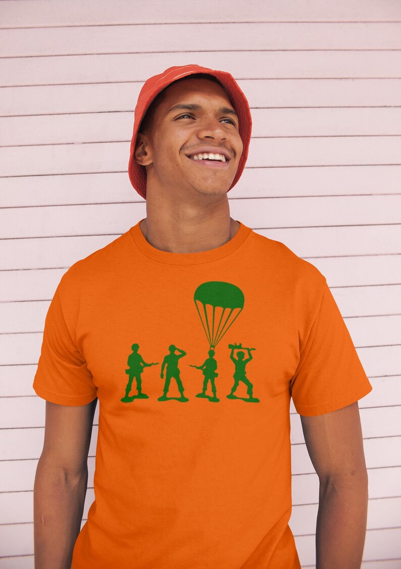 Peut inclure: Un homme portant un t-shirt orange avec une silhouette verte de quatre soldats jouets et un parachute. Les soldats tiennent des fusils jouets et le parachute est au-dessus du soldat &agrave; l'arri&egrave;re.