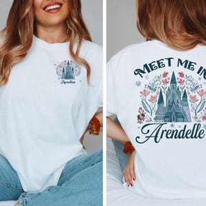 May include: White t-shirt featuring a castle design with the text "Meet Me In Arendelle" on the back. The front of the shirt has a smaller castle design and the name "Amelia". The design includes pink and blue floral elements.