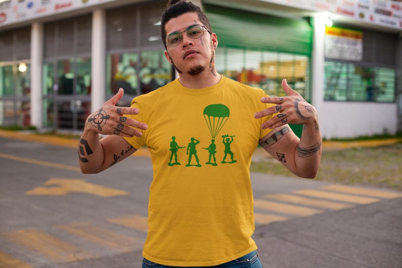 Peut inclure: Un t-shirt jaune avec un motif vert de soldats de jouets en parachute. Les soldats tiennent des fusils et un parachute est au-dessus d'eux.