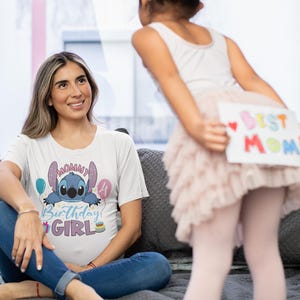 May include: A woman wearing a white t-shirt with a Stitch graphic and the text "Mommy Birthday Girl" sits on a gray bed. A child holds a sign that says "Best Mom". The scene is indoors.