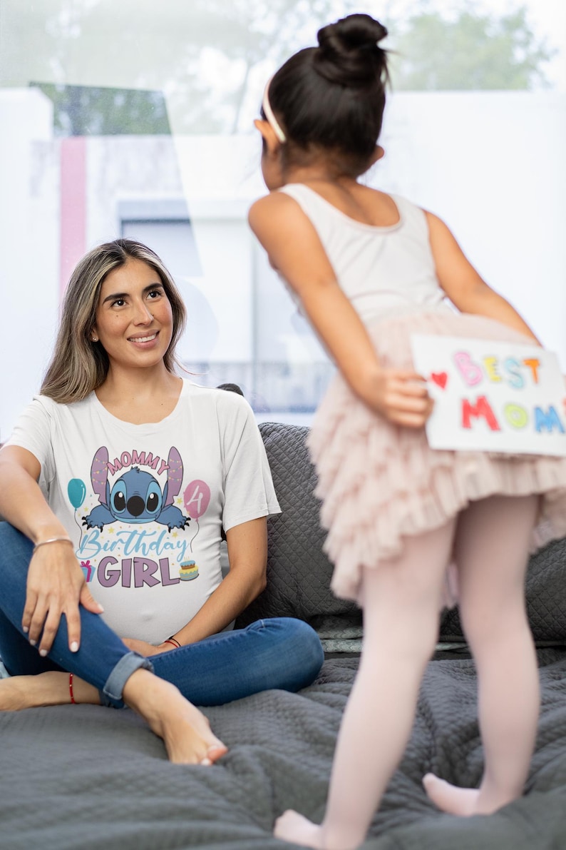May include: A woman wearing a white t-shirt with a Stitch graphic and the text "Mommy Birthday Girl" sits on a gray bed. A child holds a sign that says "Best Mom". The scene is indoors.
