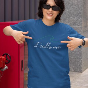 May include: A woman wearing a blue t-shirt with a green wave graphic and the text "it calls me". She is pointing at the graphic on the shirt.