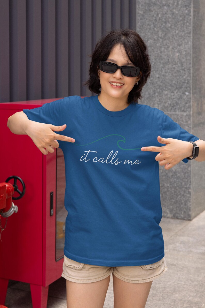 May include: A woman wearing a blue t-shirt with a green wave graphic and the text "it calls me". She is pointing at the graphic on the shirt.