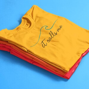 May include: A stack of folded t-shirts in yellow, orange, and red. The top shirt is yellow and has the text "it calls me" with a blue wave graphic.