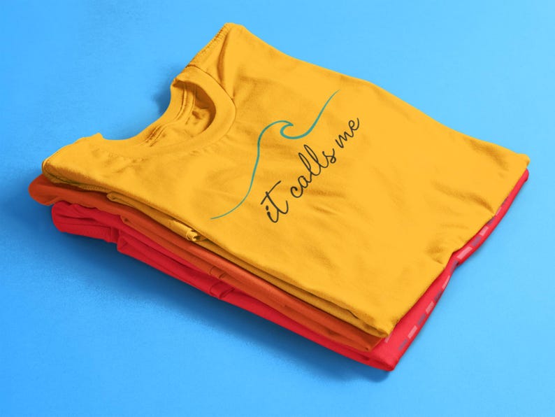 May include: A stack of folded t-shirts in yellow, orange, and red. The top shirt is yellow and has the text "it calls me" with a blue wave graphic.