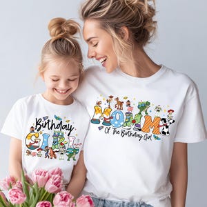 May include: White t-shirts with colorful graphics. One shirt says "Birthday Girl" with cartoon characters, and the other says "Mommy Of The Birthday Girl". The shirts are worn by a mother and daughter.