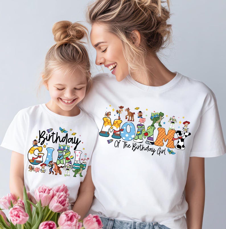 May include: White t-shirts with colorful graphics. One shirt says "Birthday Girl" with cartoon characters, and the other says "Mommy Of The Birthday Girl". The shirts are worn by a mother and daughter.
