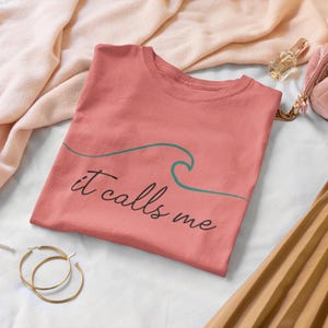 May include: A coral pink t-shirt with a green wave graphic and the text "it calls me".