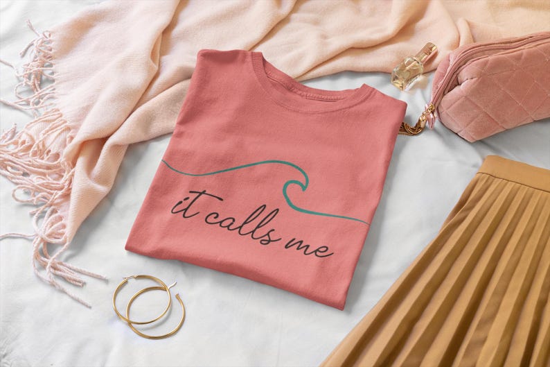 May include: A coral pink t-shirt with a green wave graphic and the text "it calls me".