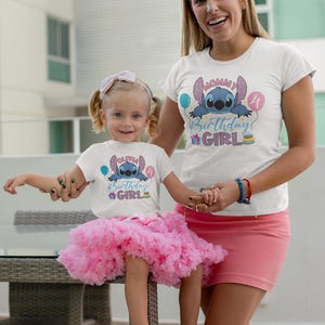 May include: Two white t-shirts with a blue cartoon character design and the text "Birthday Girl". One shirt is worn by a child wearing a pink tutu. The other shirt is worn by an adult wearing a pink skirt. The child is sitting on a table.