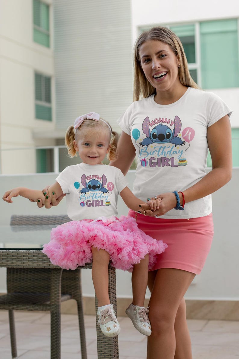 May include: Two white t-shirts with a blue cartoon character design and the text "Birthday Girl". One shirt is worn by a child wearing a pink tutu. The other shirt is worn by an adult wearing a pink skirt. The child is sitting on a table.