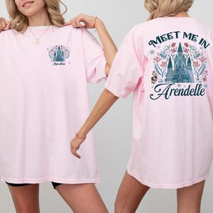 May include: Light pink t-shirts with a castle graphic. One shirt has the name "Amelia" and a castle design on the front. The other shirt has the text "Meet Me In Arendelle" with a castle graphic on the back.
