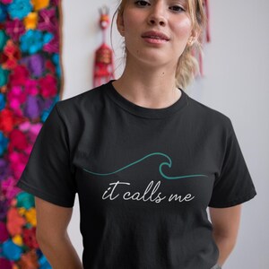 May include: A black t-shirt with a teal wave graphic and the text "it calls me" in white.