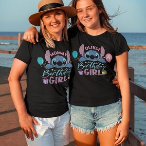 May include: Two women wearing black t-shirts with a Stitch graphic and the words "Mommy Birthday Girl" and "Olivia Birthday Girl". One woman wears a straw hat. The shirts have blue and purple accents. They are standing outside near the water.