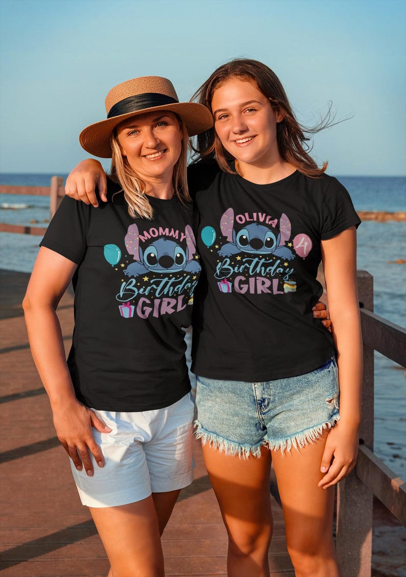 May include: Two women wearing black t-shirts with a Stitch graphic and the words "Mommy Birthday Girl" and "Olivia Birthday Girl". One woman wears a straw hat. The shirts have blue and purple accents. They are standing outside near the water.