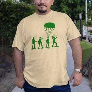 Peut inclure: Un t-shirt beige avec un motif vert d'un parachute et de quatre soldats en plastique. Les soldats sont en ligne, celui du fond tenant un parachute.