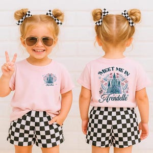 May include: A young child wearing a light pink t-shirt and black and white checkered shorts. The front of the shirt has a castle graphic and the name "Amelia". The back of the shirt says "Meet Me In Arendelle" with a castle graphic.