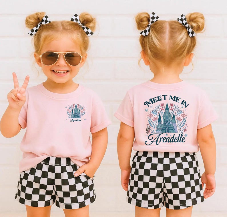 May include: A young child wearing a light pink t-shirt and black and white checkered shorts. The front of the shirt has a castle graphic and the name "Amelia". The back of the shirt says "Meet Me In Arendelle" with a castle graphic.