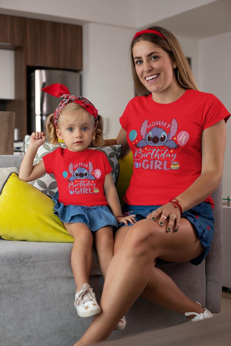 May include: Matching red t-shirts with a cartoon character and the words "Mommy Birthday Girl" and "Olivia Birthday Girl". The shirts are worn by a woman and a young girl, both wearing red headbands and denim skirts. The girl is sitting on a gray couch.