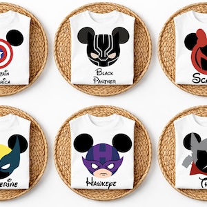 May include: White t-shirts with superhero-themed designs, each featuring a Mickey Mouse head silhouette. Designs include Captain America, Black Panther, Scarlet, Wolverine, Hawkeye, and Thor. Each shirt is displayed on a round woven placemat.