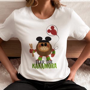 May include: White t-shirt with a cartoon image of a brown coconut monster with a Mickey Mouse hat and a red balloon. The text "Fear the Kakamora" is printed below the image.
