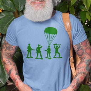 Peut inclure: Un t-shirt bleu clair avec un motif vert de quatre soldats en plastique et un parachute. Les soldats tiennent des fusils et le parachute est ouvert.