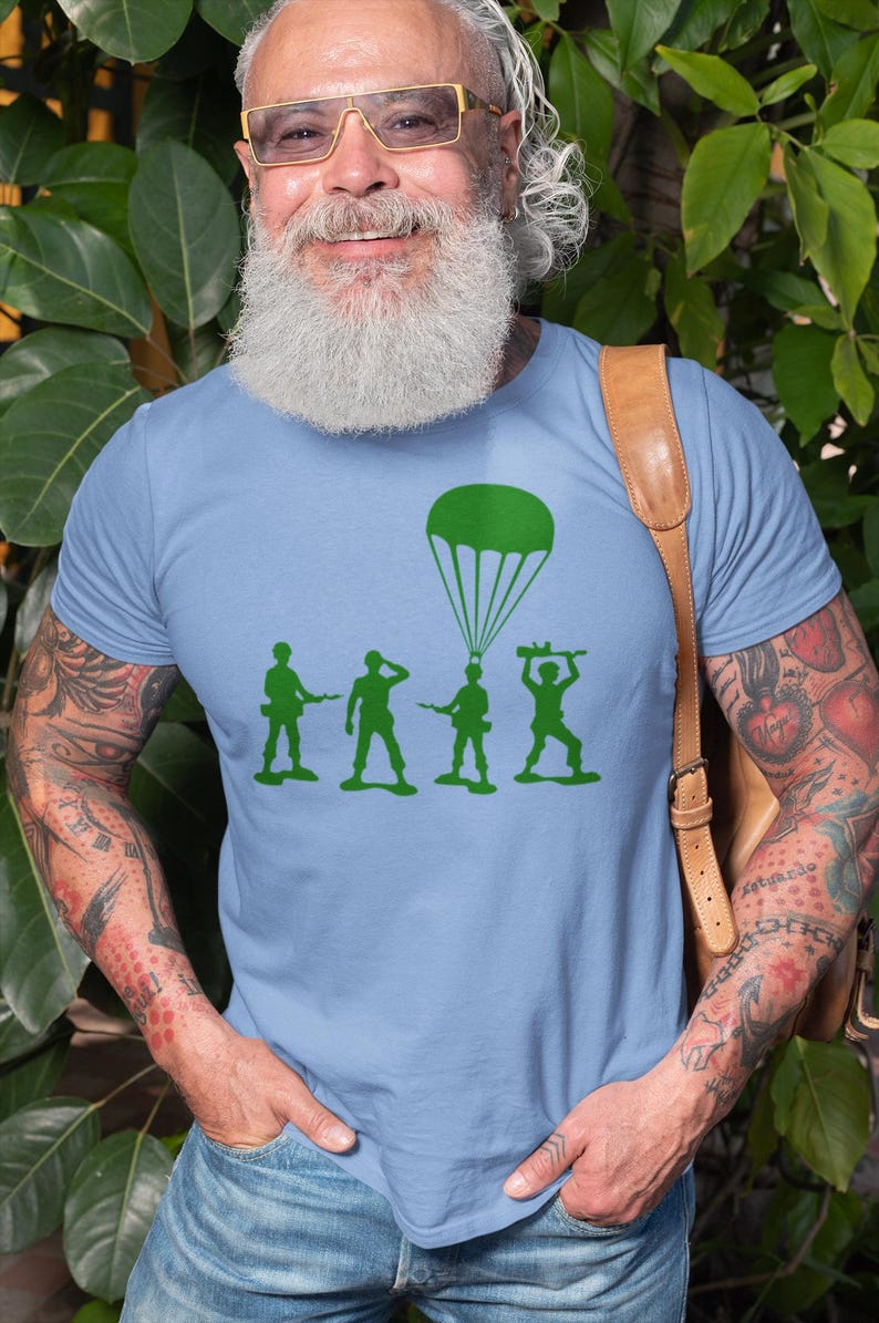 Peut inclure: Un t-shirt bleu clair avec un motif vert de quatre soldats en plastique et un parachute. Les soldats tiennent des fusils et le parachute est ouvert.