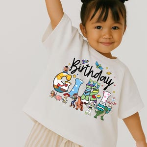 May include: A white t-shirt with the word "Birthday" in black script and the word "Girl" in colorful block letters. The letters are decorated with cartoon characters. The child is wearing beige striped shorts.