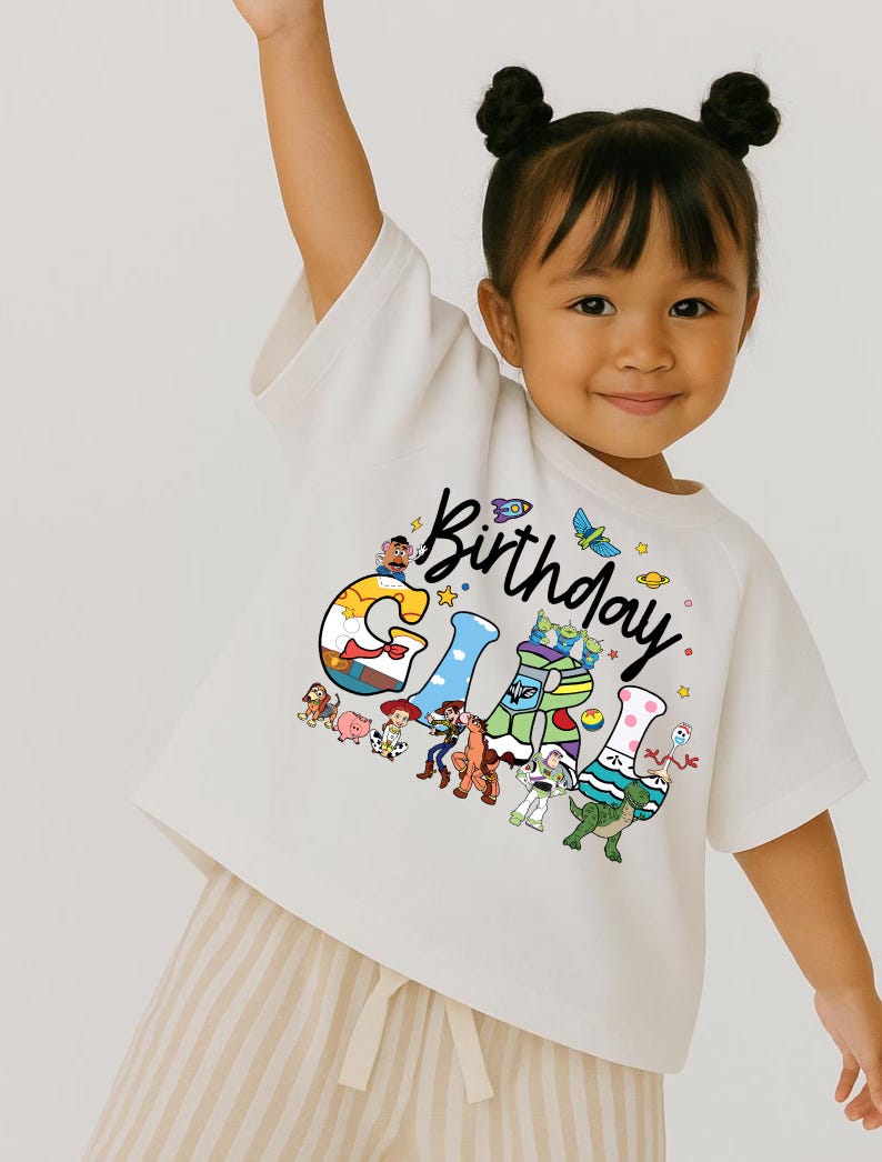 May include: A white t-shirt with the word "Birthday" in black script and the word "Girl" in colorful block letters. The letters are decorated with cartoon characters. The child is wearing beige striped shorts.