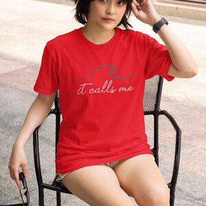 May include: A woman wearing a red t-shirt with a wave graphic and the text "it calls me". She is sitting on a black metal chair.
