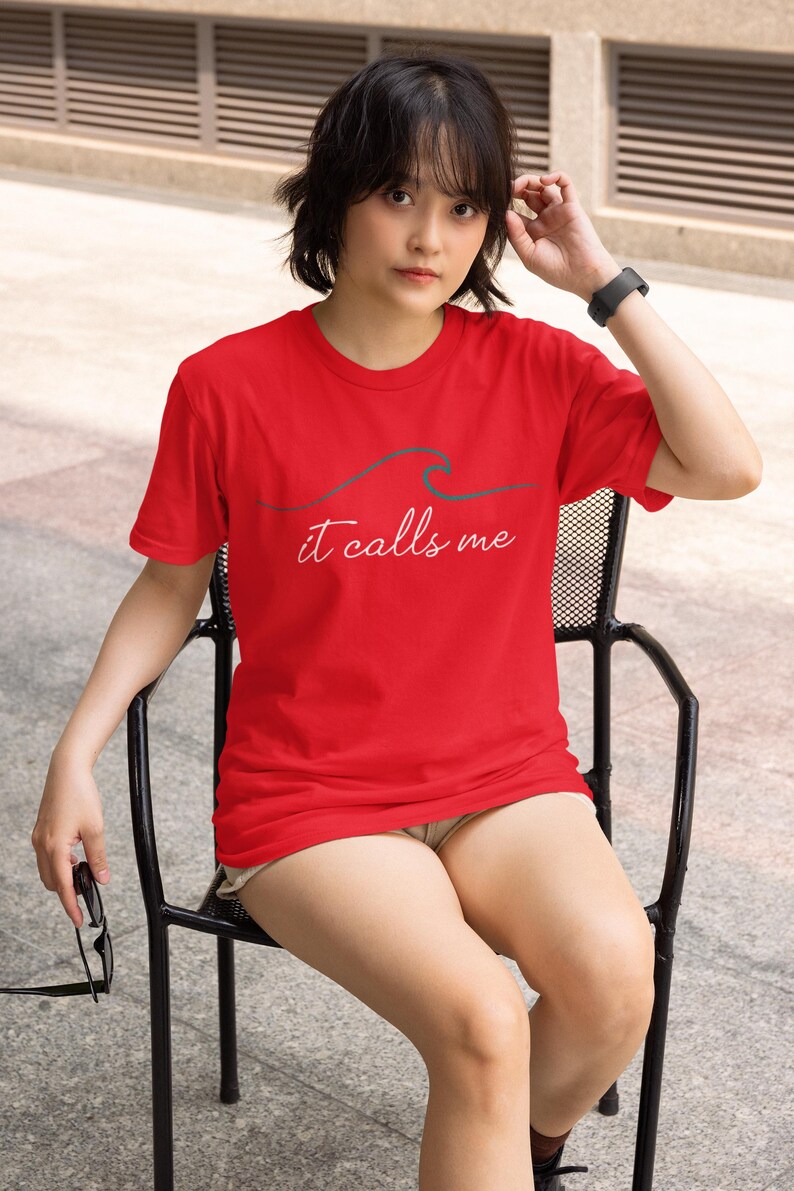 May include: A woman wearing a red t-shirt with a wave graphic and the text "it calls me". She is sitting on a black metal chair.