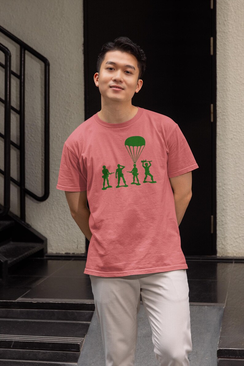 Peut inclure: Un homme portant un t-shirt rose avec un motif vert de soldats en jouet et un parachute. Le motif dit "Paratrooper".
