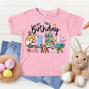 May include: Pink short-sleeved t-shirt with the words "Birthday Girl" in a playful font. The letters are decorated with cartoon characters. A denim pair of jeans, a nest with Easter eggs, and a stuffed bunny are also in the image.