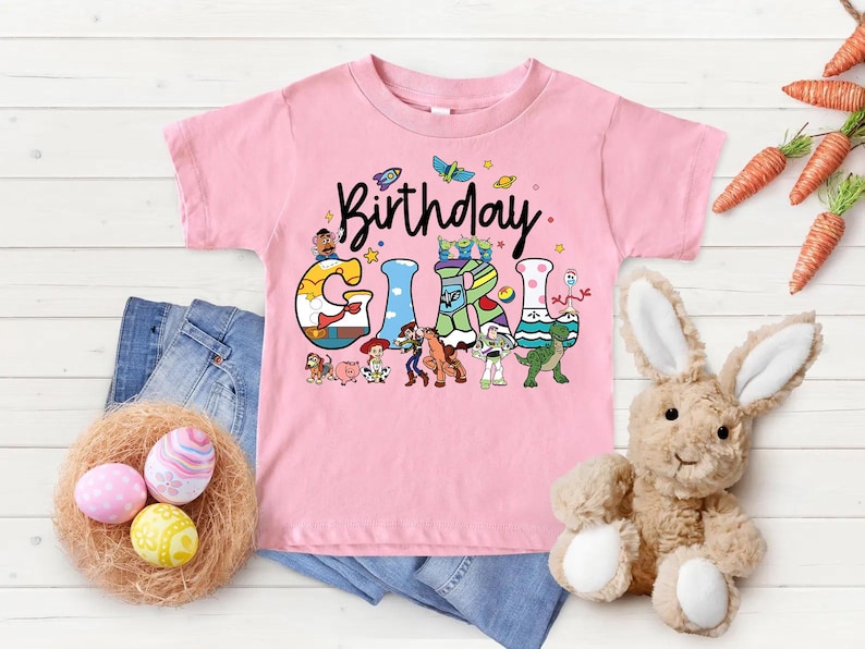May include: Pink short-sleeved t-shirt with the words "Birthday Girl" in a playful font. The letters are decorated with cartoon characters. A denim pair of jeans, a nest with Easter eggs, and a stuffed bunny are also in the image.