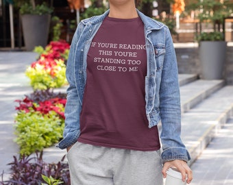 May include: A woman wearing a maroon t-shirt with the text "If you're reading this you're standing too close to me" in white. She is wearing a denim jacket and grey sweatpants. She is holding a white to-go cup.