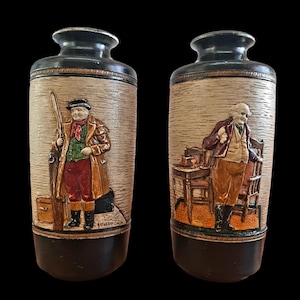 May include: Two ceramic vases with a brown and beige textured finish. Each vase features a raised relief image of a man in a period costume. One vase depicts a man holding a walking stick and a suitcase, the other depicts a man sitting in a chair. The text "Weller" is visible on the bottom of one vase.