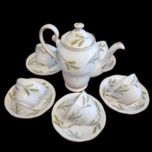 Peut inclure: Service à thé en porcelaine blanche comprenant une théière, quatre tasses et des soucoupes. L'ensemble présente un délicat motif de feuilles vertes et dorées. Les bords des tasses et des soucoupes sont dorés.