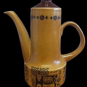 Pode incluir: Uma cafeteira de cerâmica marrom com um design floral preto e marrom e uma ilustração de um carneiro. A cafeteira tem uma tampa marrom e um bico.