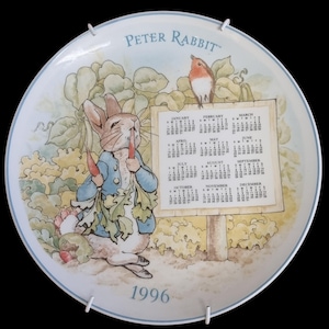 Puede incluir: Plato decorativo con Peter Rabbit comiendo zanahorias, con un calendario de 1996. Un pequeño pájaro se posa sobre el calendario. El plato tiene un borde azul y las palabras "Peter Rabbit" en la parte superior.