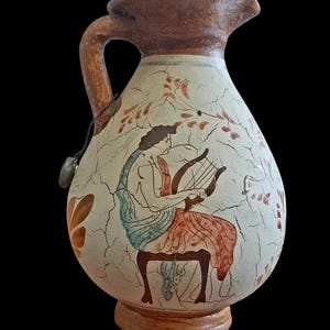May include: A ceramic pitcher with a brown handle and spout, featuring a painted scene of a figure playing a lyre. The pitcher has a cream-colored base with a cracked effect and floral accents. The base is brown.