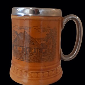Peut inclure: Une tasse en céramique brune avec un bord argenté et une illustration en noir et blanc d'une maison avec un toit de chaume. La tasse a une anse et le texte "LONDONDERRY, ALBACONNER" est imprimé au bas.