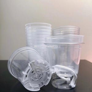 Puede incluir: Maceteros de plástico transparente con orificios de drenaje y una cuerda blanca. Las macetas están apiladas y dispuestas sobre una superficie oscura. El texto "@plantedbyaudrey" es visible.