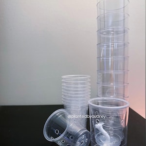 May include: Clear plastic self-watering plant pots. The pots are cylindrical with a clear design, and some have a white wick. The pots are stacked and arranged on a black surface. The text "@plantedbyaudrey" is visible.