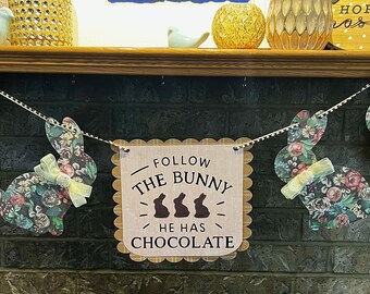 May include: A brown banner with scalloped edges and the text "Follow the bunny he has chocolate" with three chocolate bunny silhouettes. The banner is hanging from a string with floral patterned bunny cutouts.