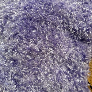 May include: A close-up view of a light purple fabric with a dense, curly, and shaggy texture. The material features a consistent lavender hue throughout, with individual fibers creating a looped, high-pile surface that appears soft and voluminous.