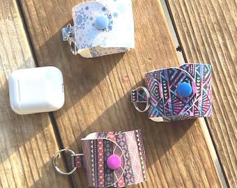 May include: Four different AirPod cases with keychains. One case is white with a snowflake pattern, one is blue and pink with a geometric pattern, and two are pink and orange with a geometric pattern. All cases have a button on the side.
