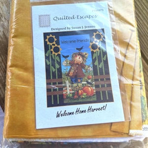 Könnte beinhalten: Ein verpacktes Quilt-Set, entworfen von Susan J. Jensen. Das Set enthält Stoff und ein Muster mit einem Herbstthema, einschließlich einer Vogelscheuche, Kürbissen und Sonnenblumen. Der Text auf dem Muster lautet "Welcome Friends" und "Welcome Home Harvest!"