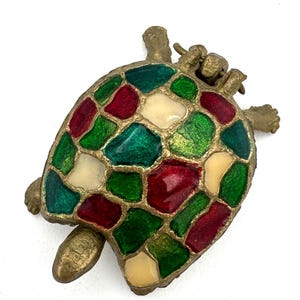 May include: A decorative turtle-shaped brooch with a mosaic design. The brooch features a gold-colored metal frame and a shell composed of red, green, and cream-colored glass pieces. The turtle has a detailed head, legs, and tail.